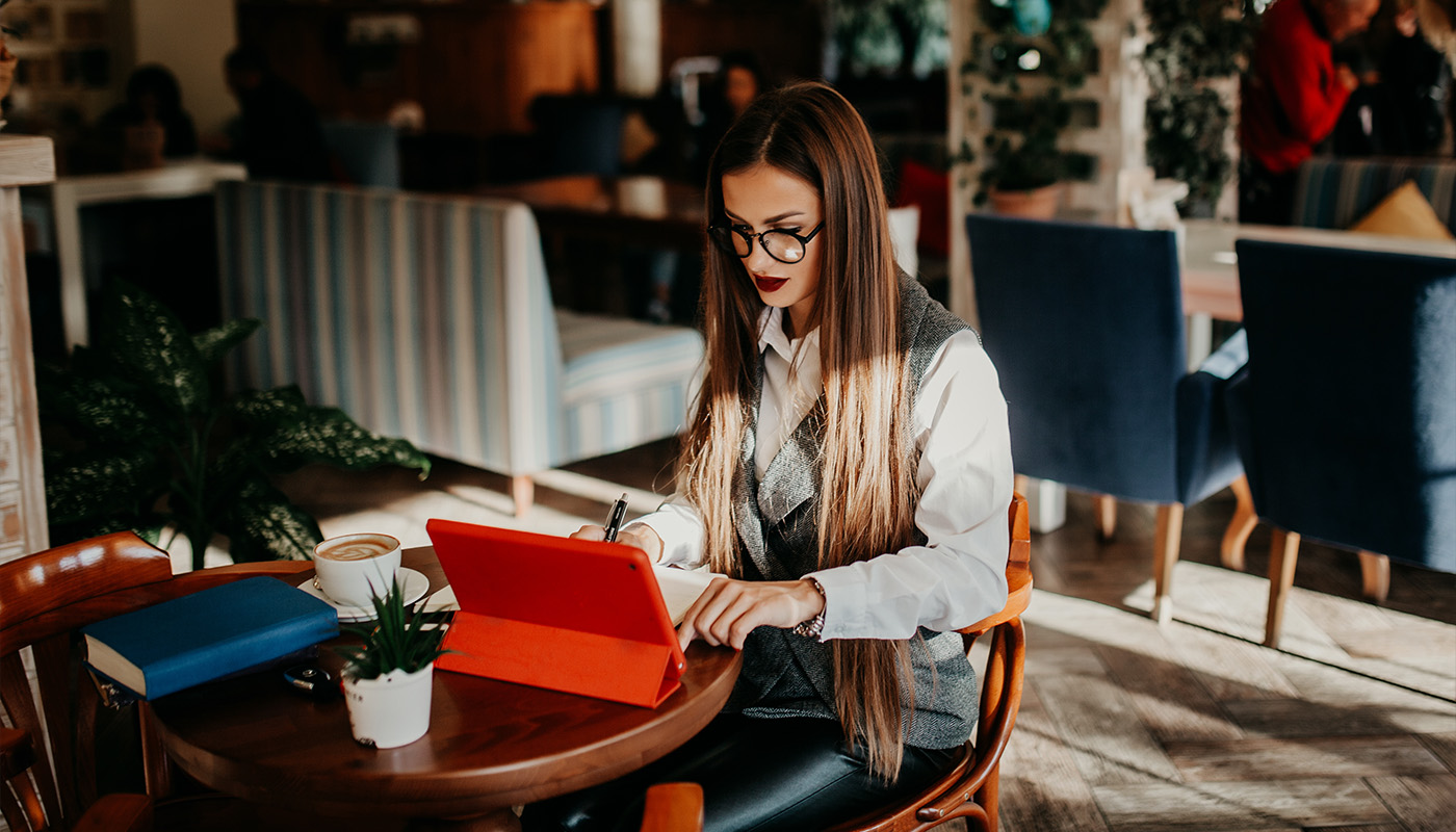 Young woman at a café with a red tablet, coffee, blue book, and succulent, reflecting an inviting workspace for C&D Advisory.